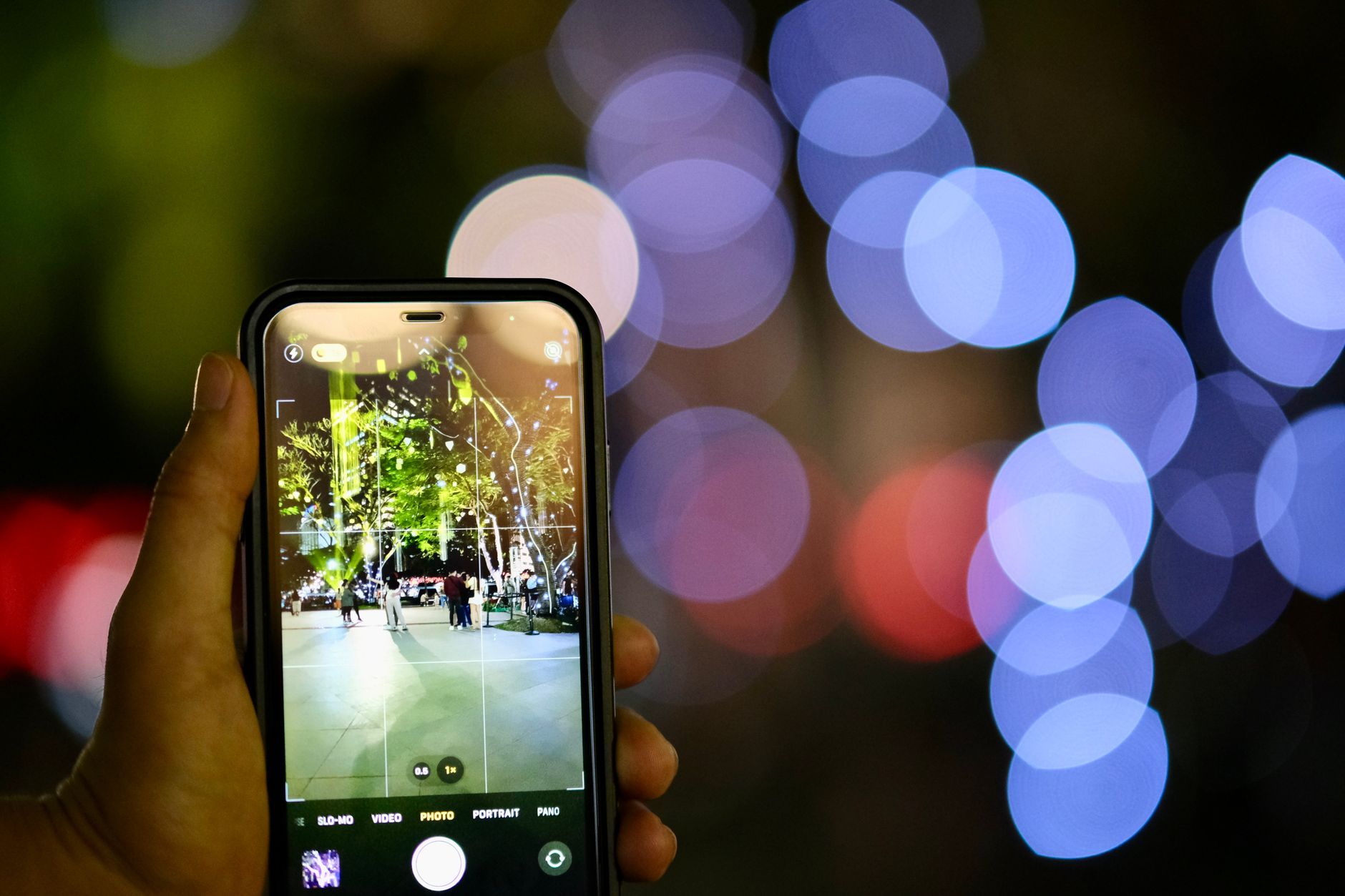 A close-up of a hand holding a smartphone capturing a night scene with bokeh lights.