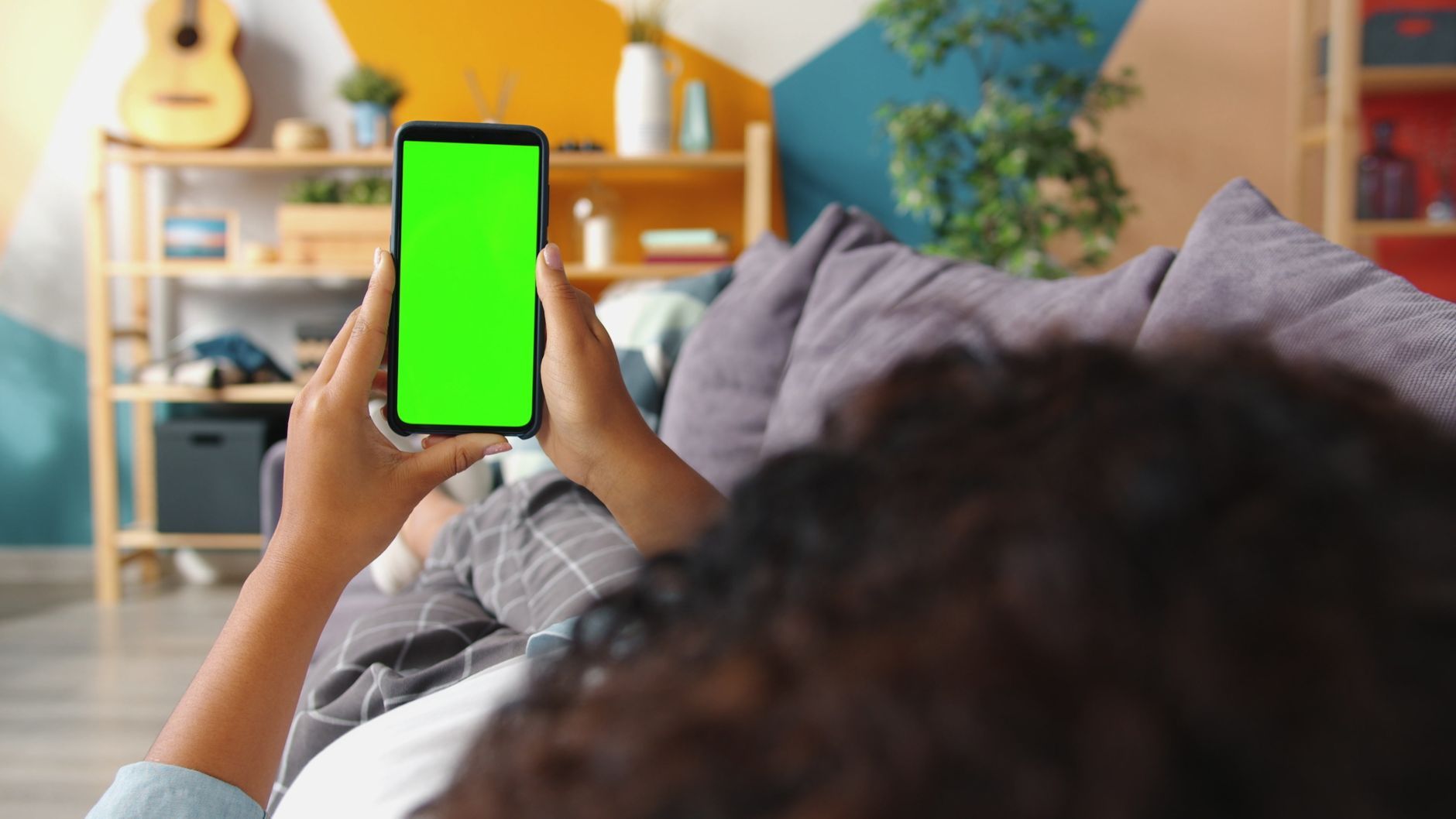 Person holding smartphone with green screen while relaxing on a couch indoors.