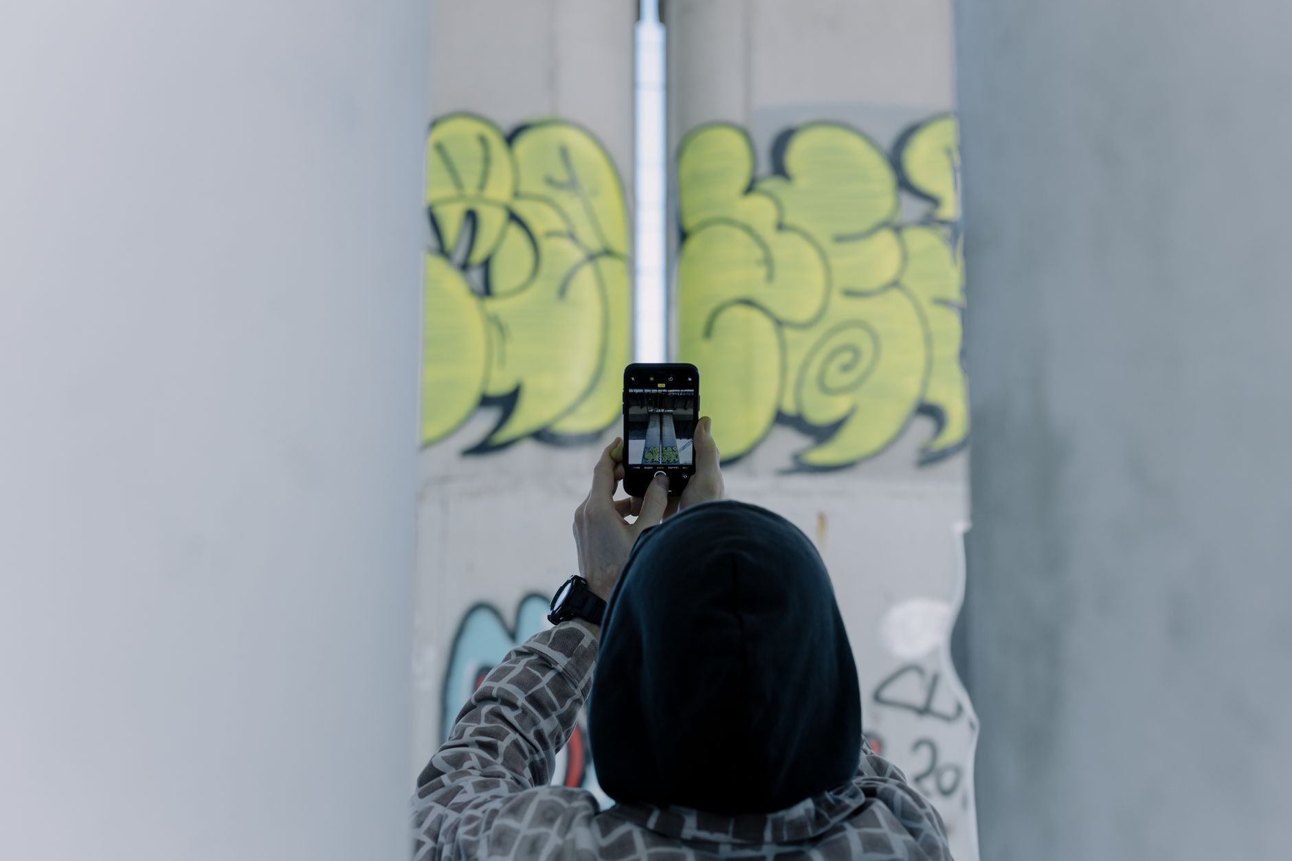 A person in a hoodie photographing urban graffiti art using a smartphone outdoors.